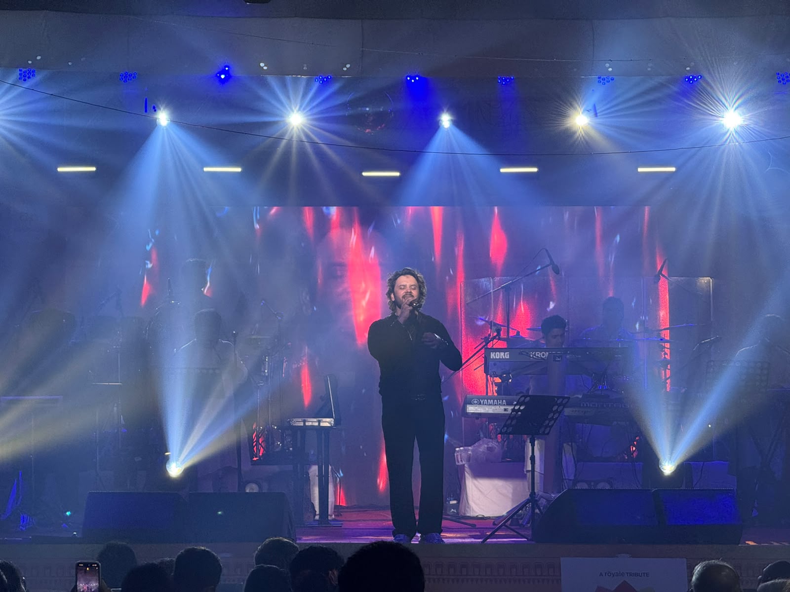 Male vocalist with live band performing under intense white and blue beam lighting at a Royale tribute concert