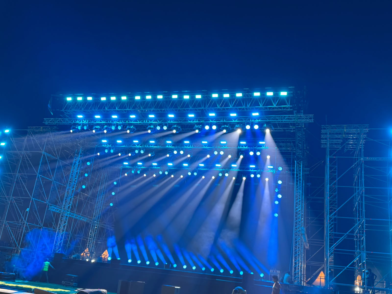 Outdoor festival stage bathed in deep blue lighting with dense beam rig and green top-lit truss