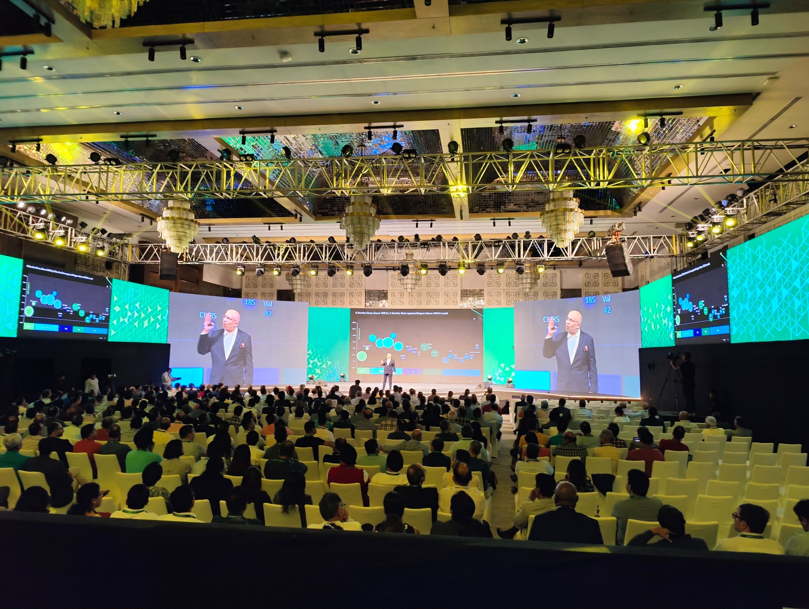 Corporate keynote on a grand stage with teal LED walls, truss lighting and packed ballroom audience