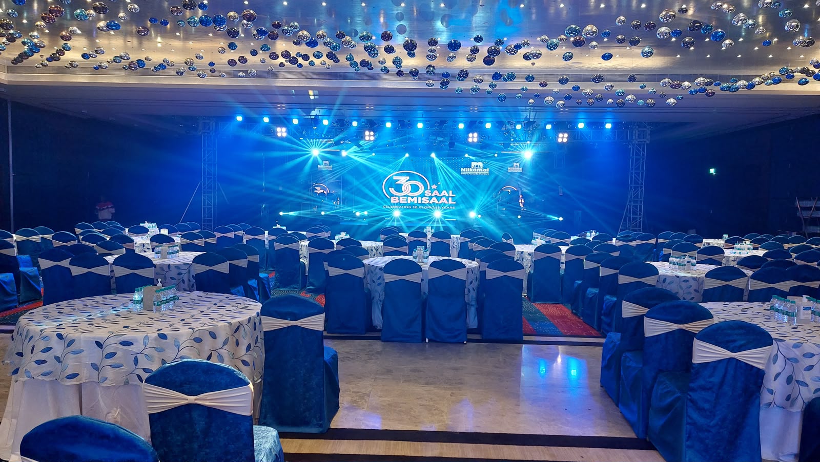 Nilkamal 30 Saal Bemisaal banquet room set with blue chair covers and lit stage