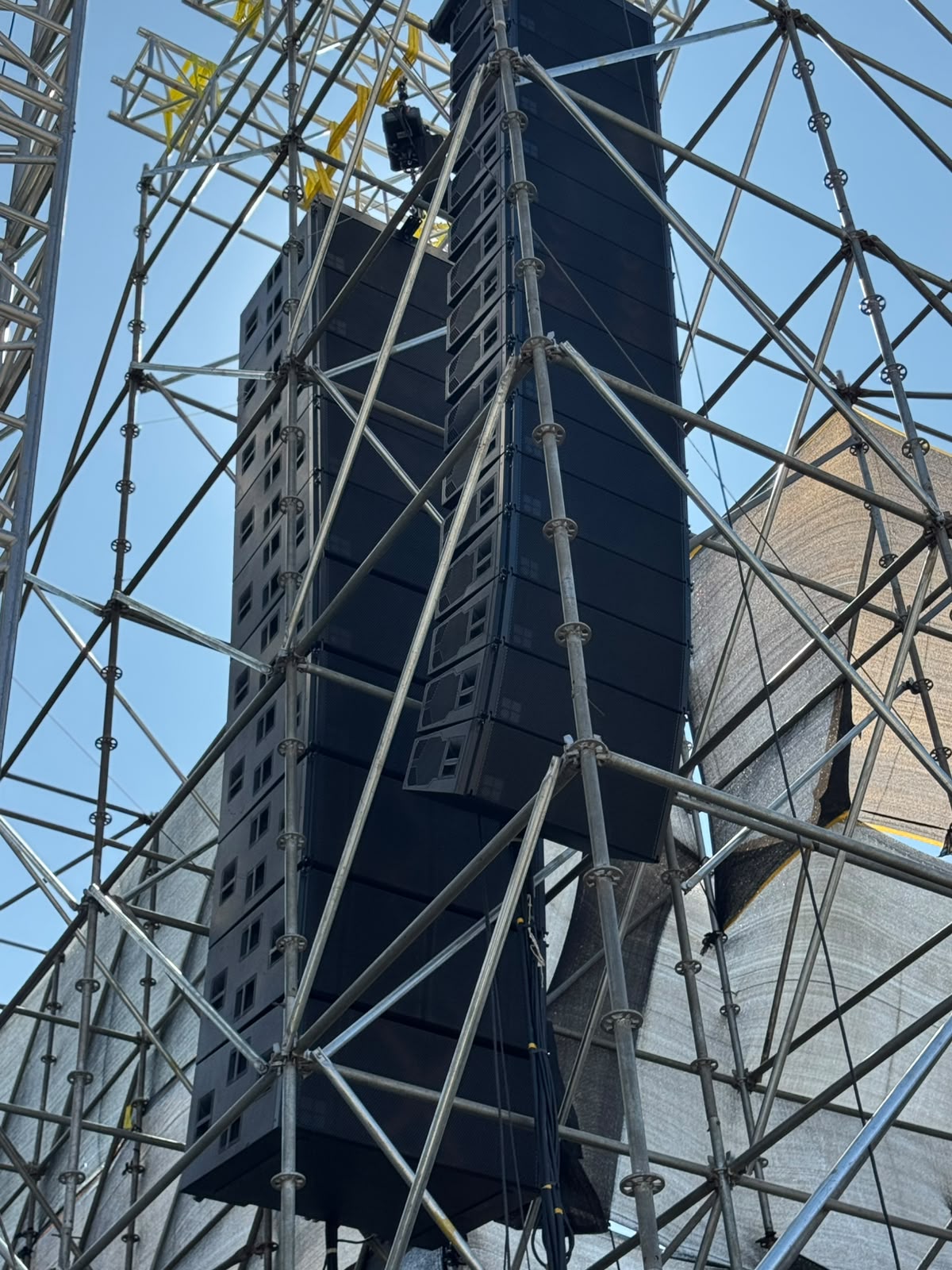 Line array speaker stack hung from scaffold tower against a clear blue sky at a festival build