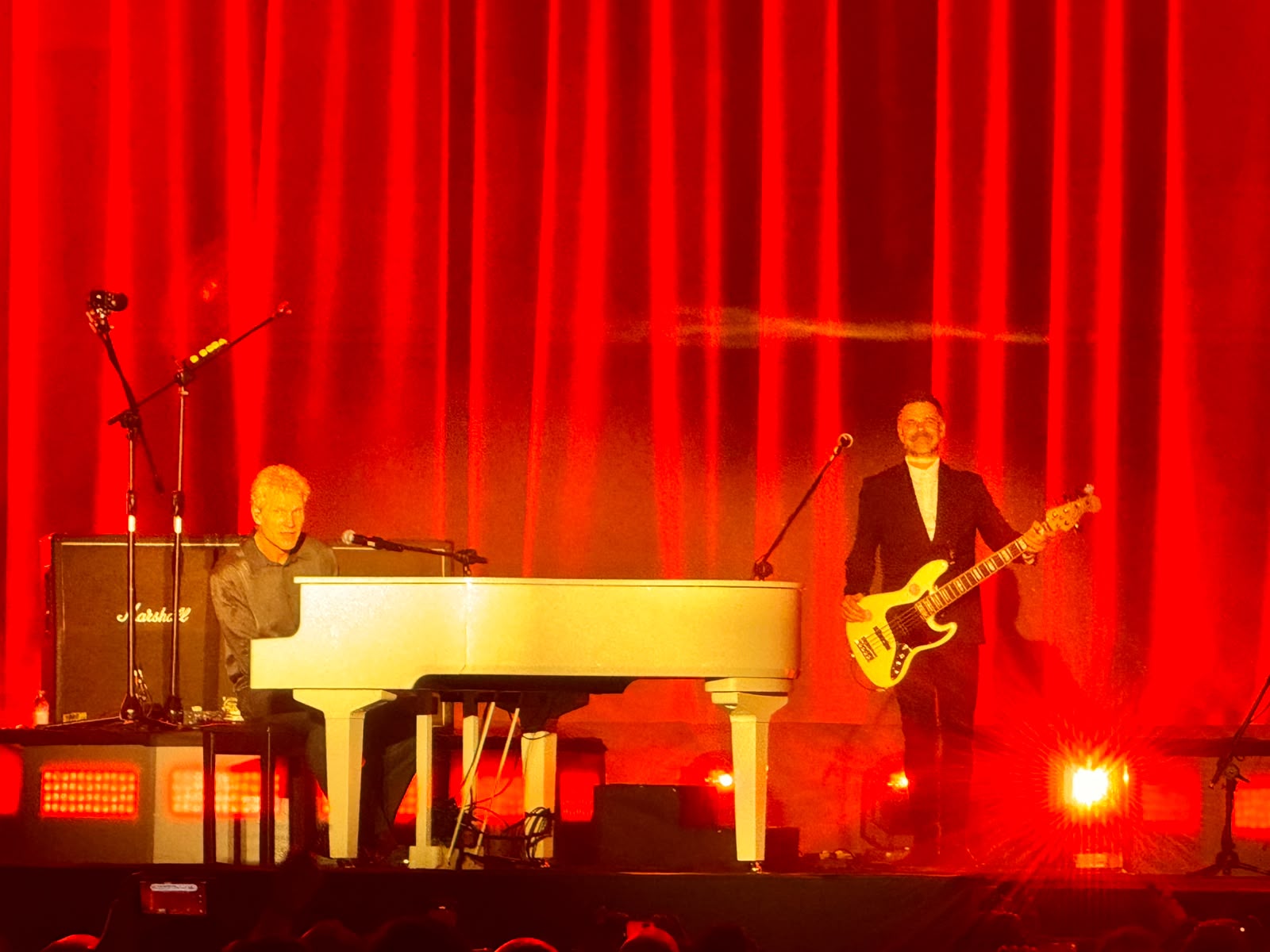 Michael Learns to Rock lead singer at a white grand piano with bassist beside him against a red curtain backdrop during their 2025 India Tour