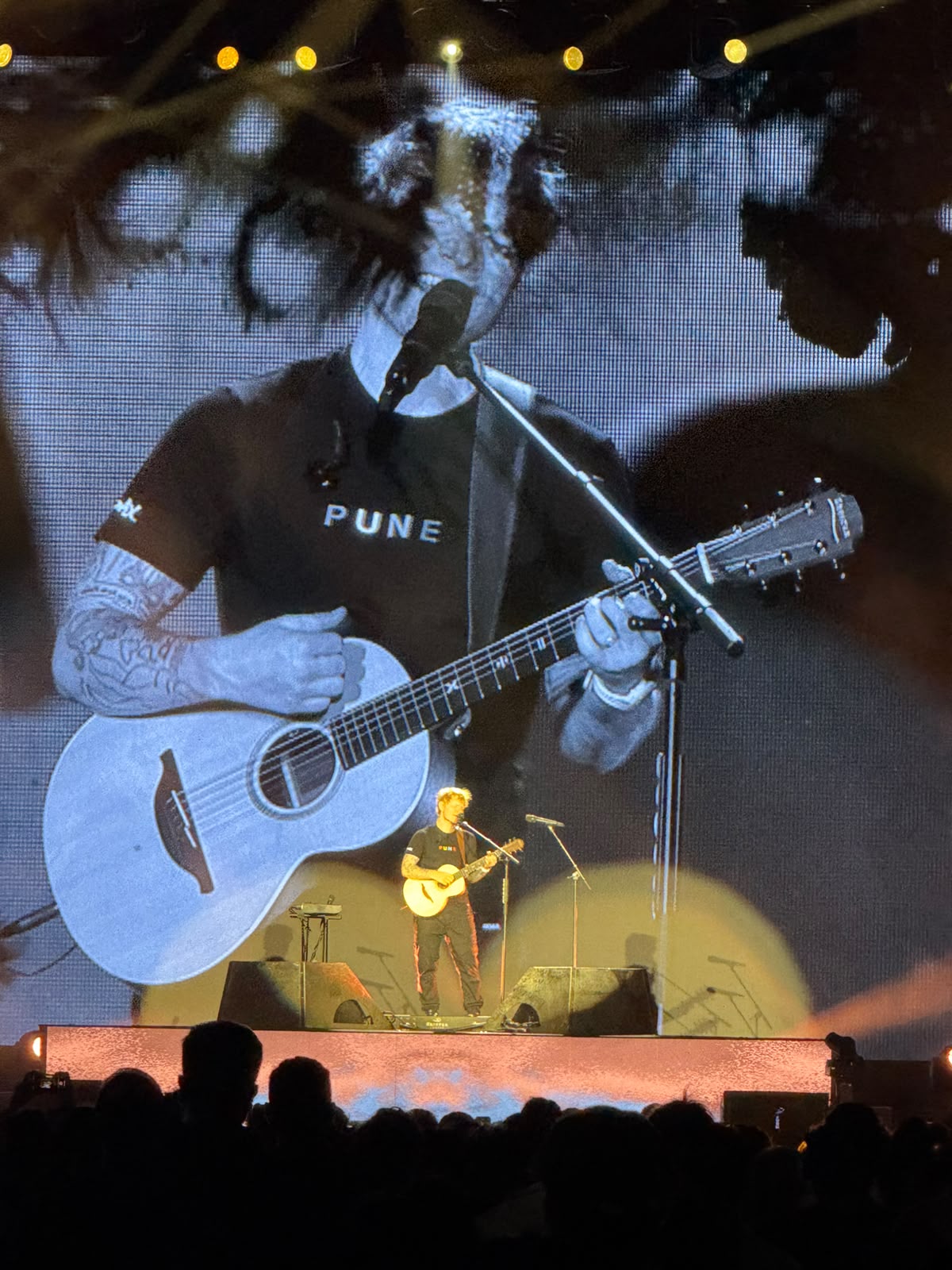 Ed Sheeran performing solo with acoustic guitar in front of a massive LED screen showing his close-up during the Pune show of his 2025 India Tour